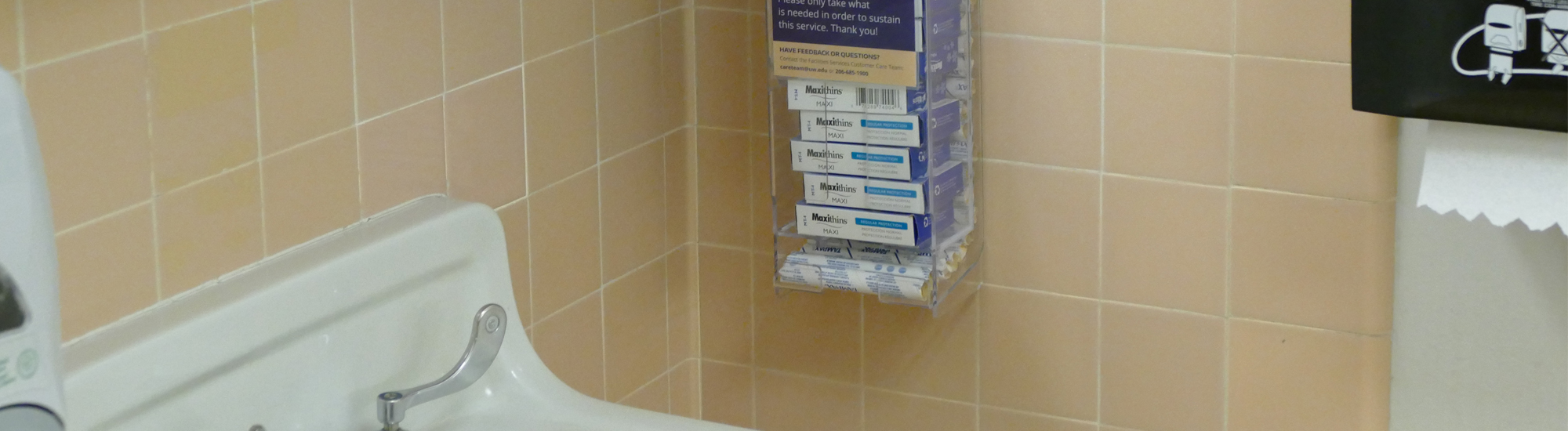 A clear acrylic dispenser affixed to a tiled wall holds individually-wrapped tampons and pads