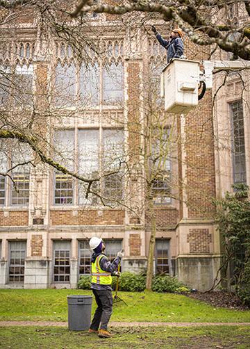 man in cherry picker points out something to a man on the ground raking branches