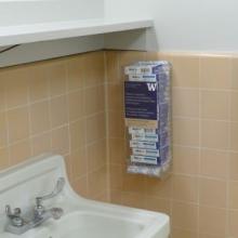Dispenser container stocked with feminine products mounted to tiled wall in restroom