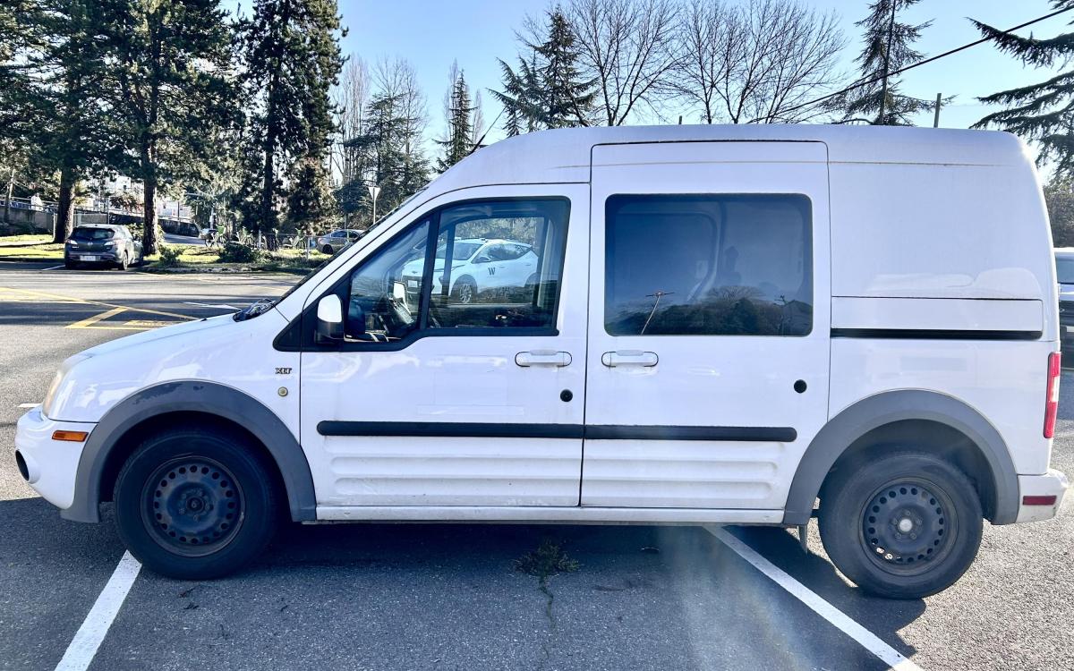 White Ford Transit in a parking lot with trees and blue sky behind.
